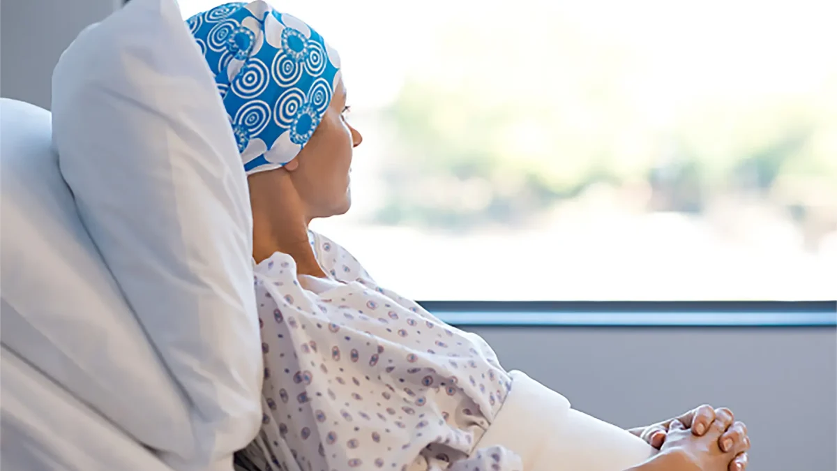 Woman in hospital gown and head covering sits by window, looking out.