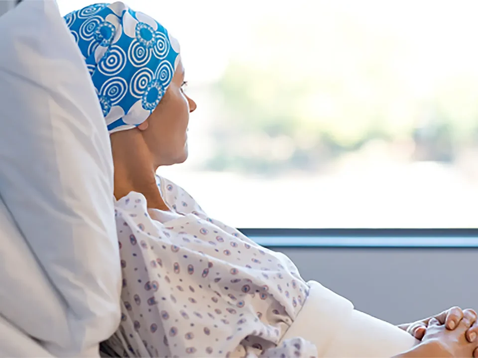 Woman in hospital gown and head covering sits by window, looking out.